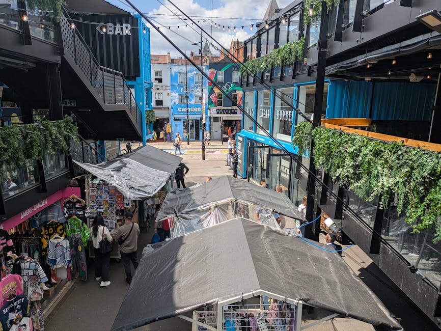 Exterior view of Camden Market showcasing a row of vibrant, multi-story shopfronts under a cloudy sky. The shops feature various signage, including a large 'COLD STEEL' sign on a brick building and de