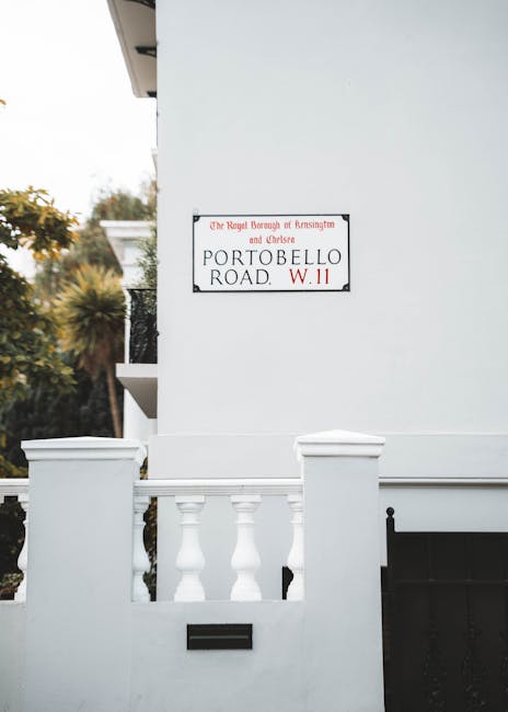 Photograph of a white exterior wall featuring a street sign reading 'Portobello Road, W11' mounted on the wall of a building in London. The building has a decorative white balustrade with classical-st