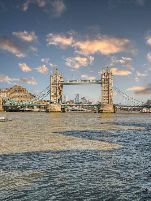 A view of Tower Bridge in London during daytime with a partly cloudy sky. The bridge's twin towers, made of stone and featuring Gothic Revival architecture, are connected by a central span that lifts