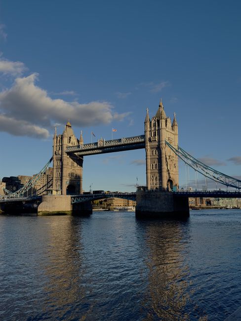 A view of Tower Bridge in London during daytime with a partly cloudy sky. The bridge's twin towers, made of stone and featuring Gothic Revival architecture, are connected by a central span that lifts